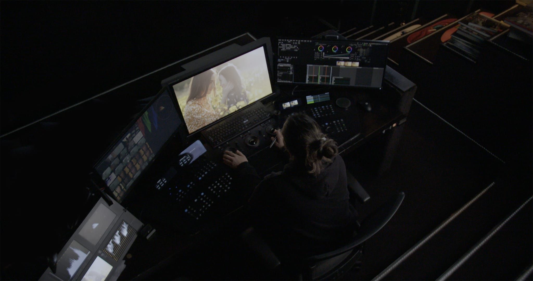 Colorist at a grading desk seen from above, with the client monitor and scopes lit in a dark suite